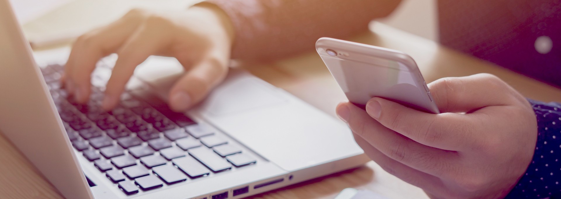 Person sitzt am Laptop und hat Ihr Handy in der Hand
