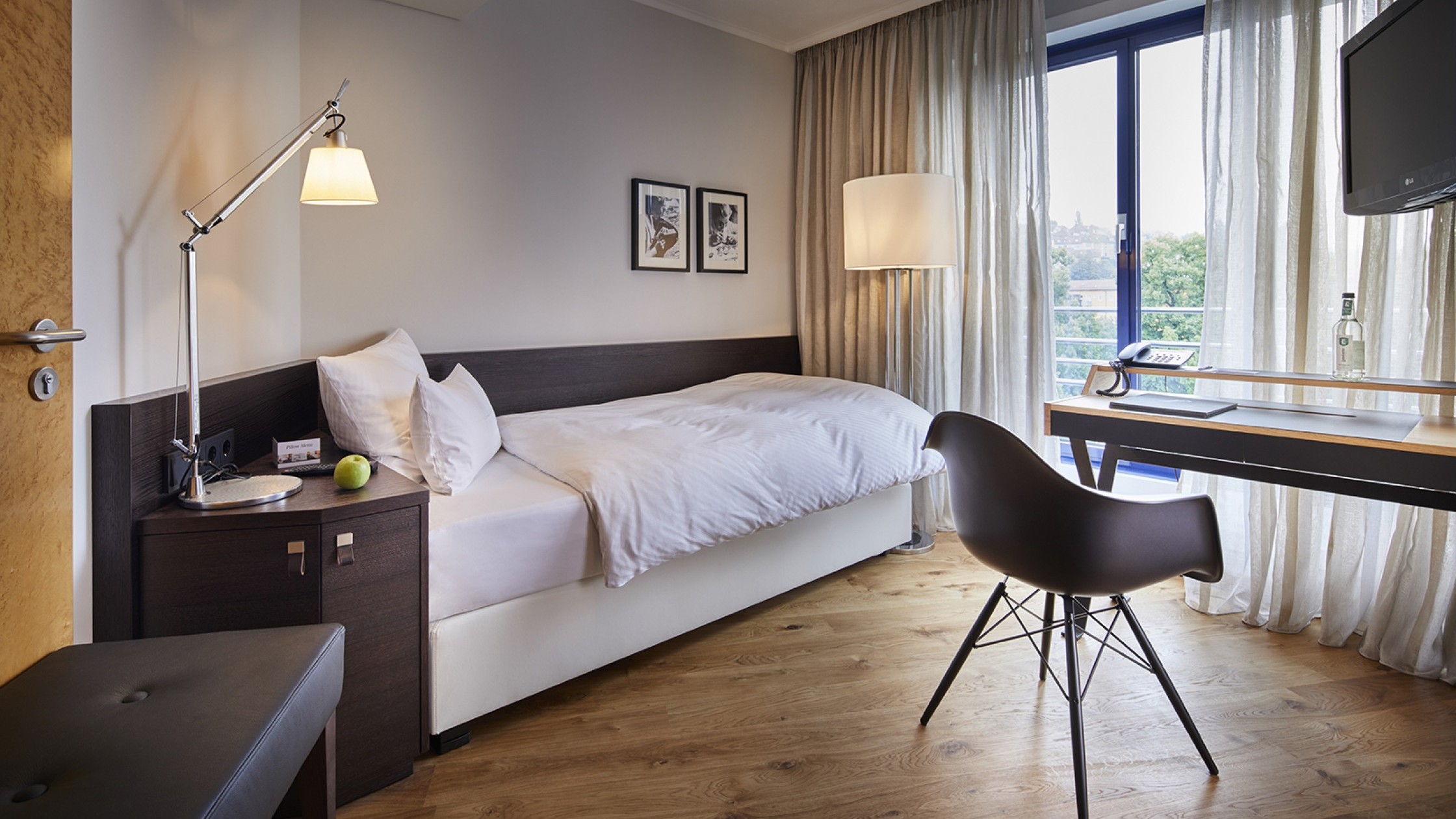 Blick ins Einzelzimmer auf das Einzelbett. Rechts im Raum steht ein Schreibtisch von dem aus man durch das große Fenster Blick auf den Park hat