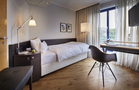 Blick ins Einzelzimmer auf das Einzelbett. Rechts im Raum steht ein Schreibtisch von dem aus man durch das große Fenster Blick auf den Park hat