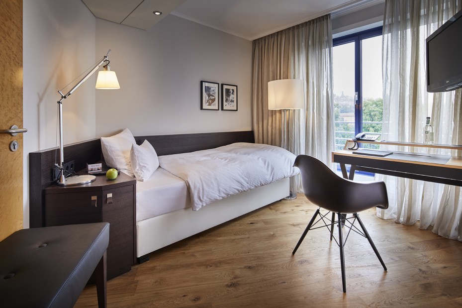 Blick ins Einzelzimmer auf das Einzelbett. Rechts im Raum steht ein Schreibtisch von dem aus man durch das große Fenster Blick auf den Park hat