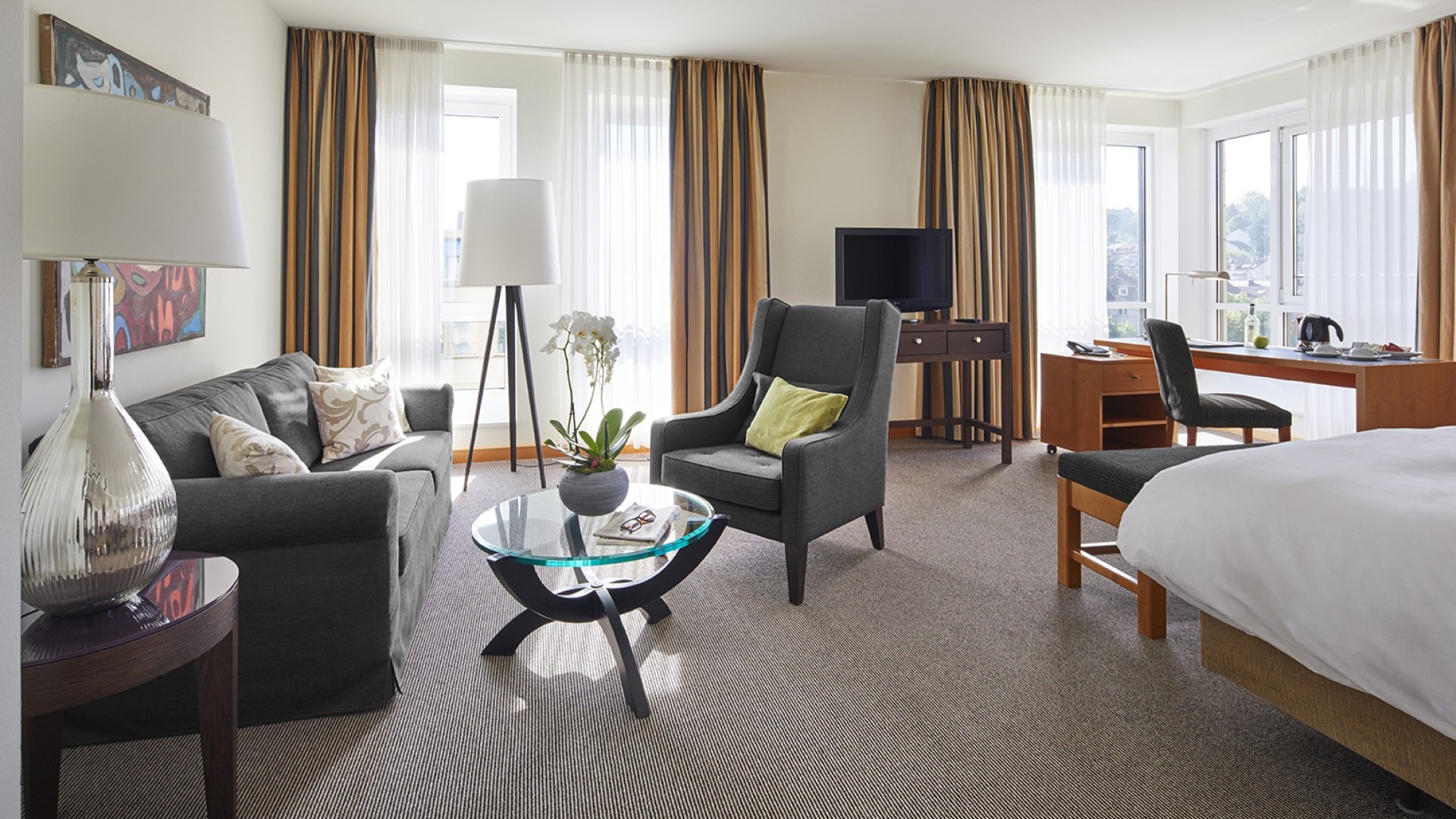 Wohnbereich mit Sofa und Sessel, im Hintergrund Fernseher und Schreibtisch vor den Fenstern